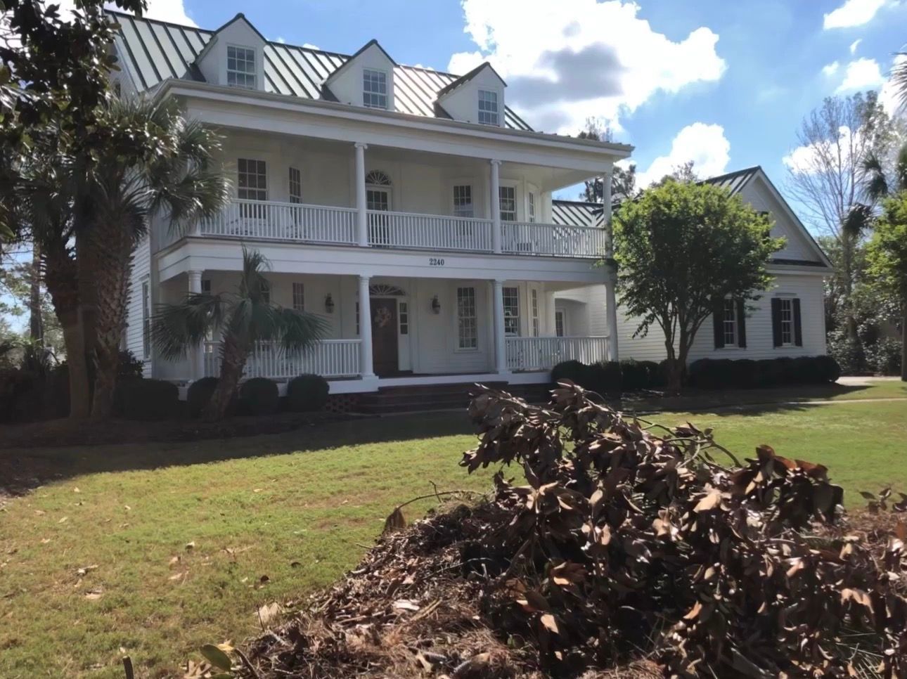 White two-story house with a porch and dormers, set on a green lawn with a pile of brown leaves.