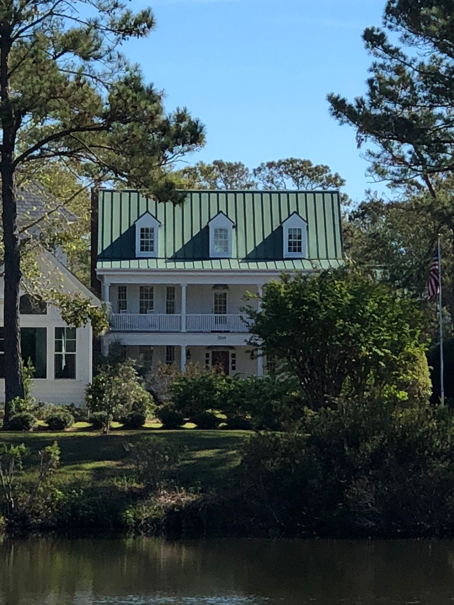 Two-story white house with green roof and dormer windows, surrounded by trees and a pond.