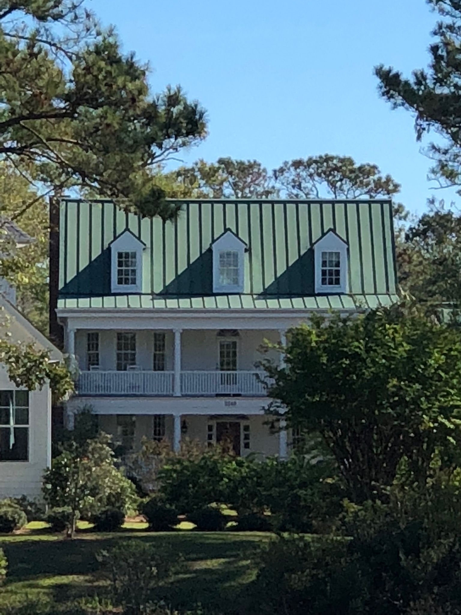 Two-story white house with green metal roof, three dormer windows, and a porch, set amongst trees.