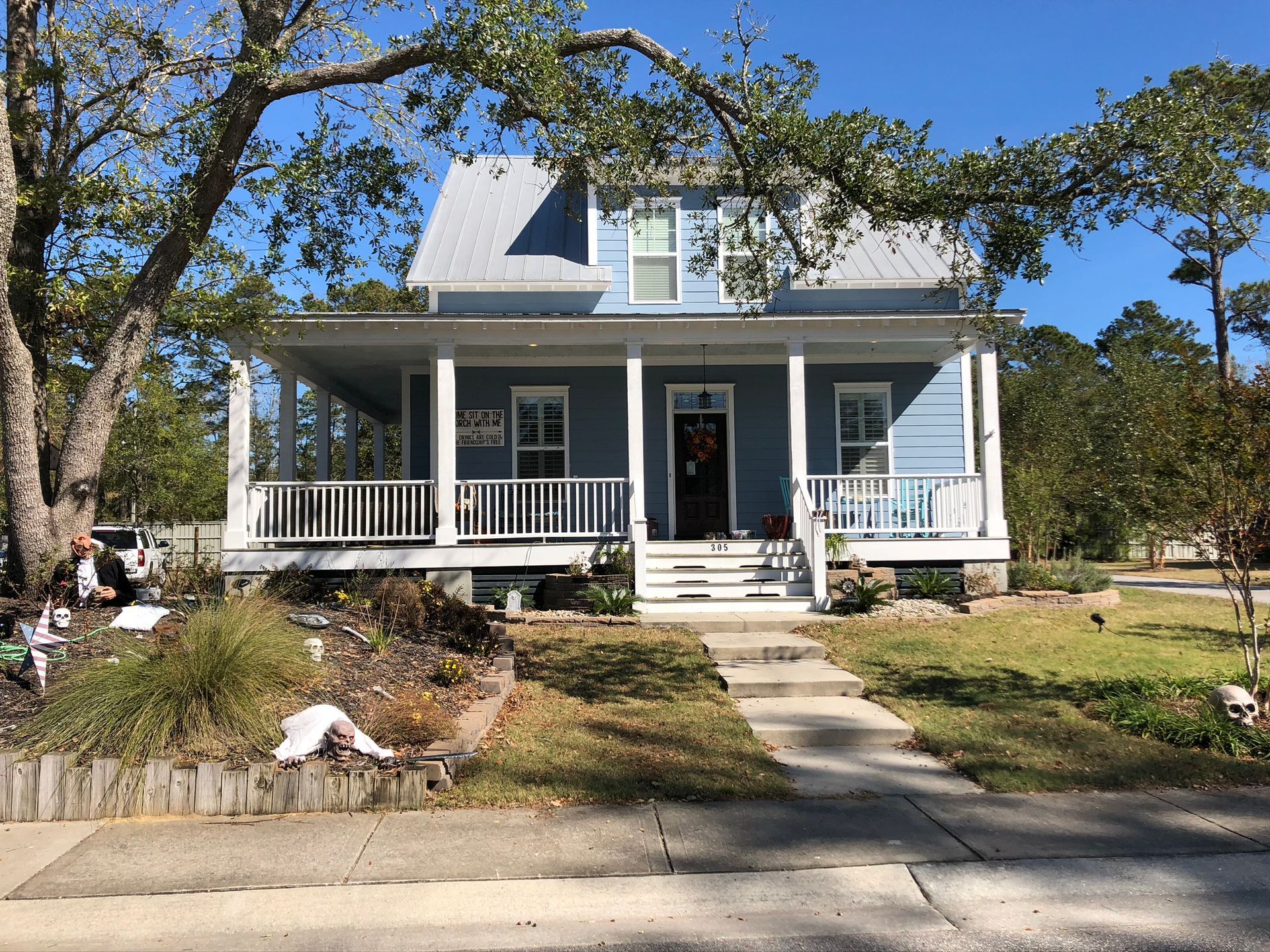 Blue house with white porch and metal roof, under a large tree.
