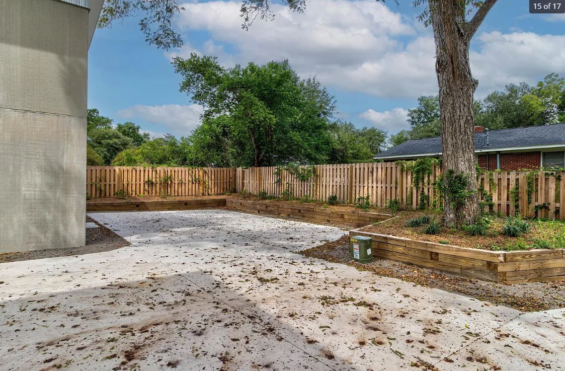Backyard with unfinished concrete patio, wooden fence, tree, and vegetation. Cloudy sky in background.