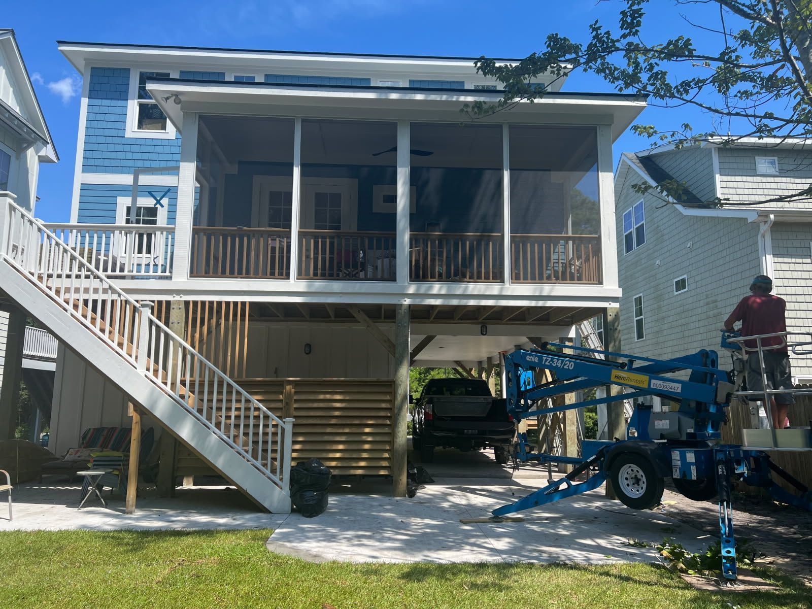 Two-story beach house with screened porch. A person on a lift works on the exterior. Driveway underneath.