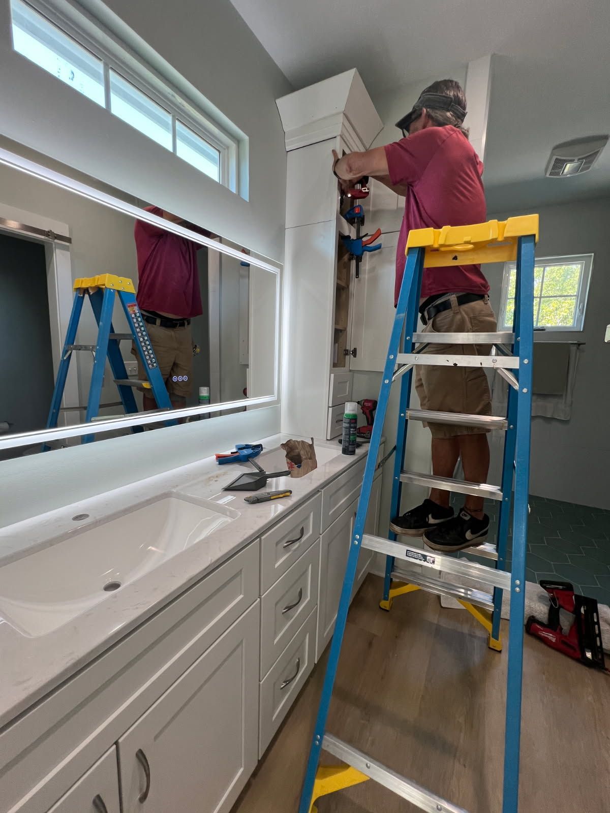 Person on ladder working on cabinet in bathroom. White cabinets, large mirror, and tools are visible.
