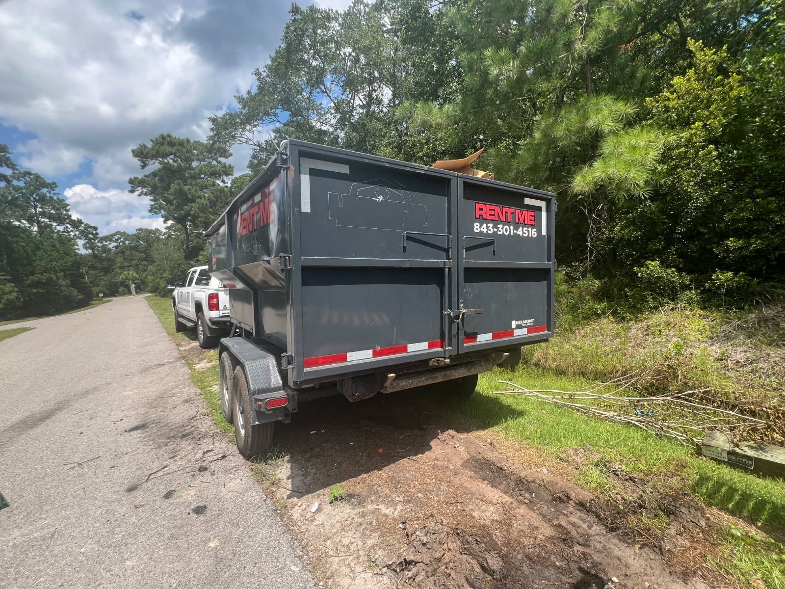 A dark gray dumpster trailer parked on the side of a road, with a white truck.