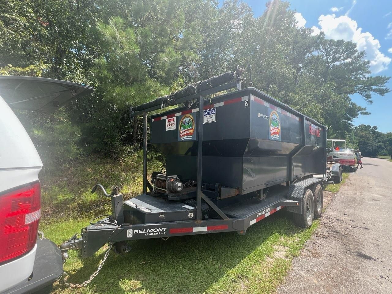 Black dumpster on trailer hitched to a white vehicle, parked on a grassy verge, trees in the background.