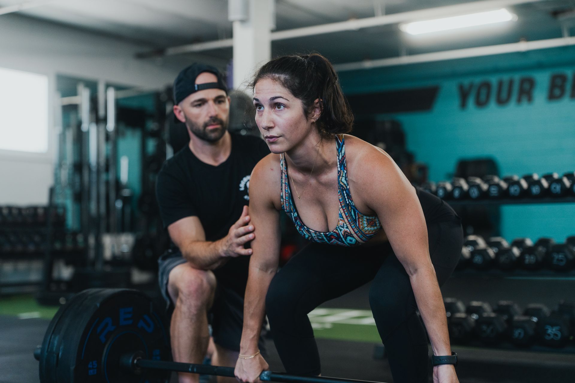 A man is lifting a barbell in a gym while another man watches.