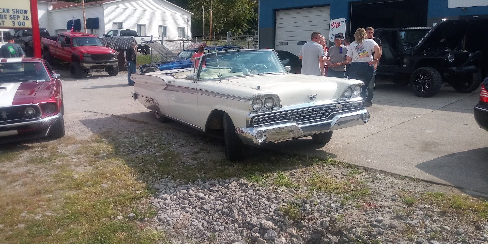 A white vintage convertible parked in a gravel lot with a crowd of people gathered around other cars on a sunny day.
