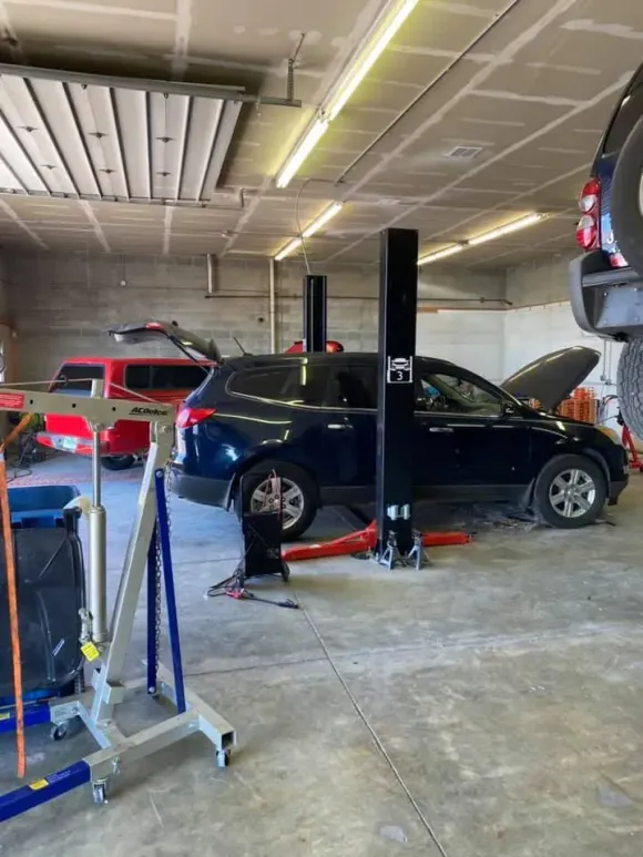 Two cars in an auto repair shop, one black SUV lifted on a jack with its hood open and a red truck nearby.
