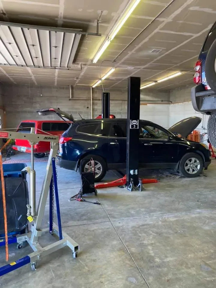 Car repair shop with a blue SUV on a lift and a red truck in the background; hood open on another car.