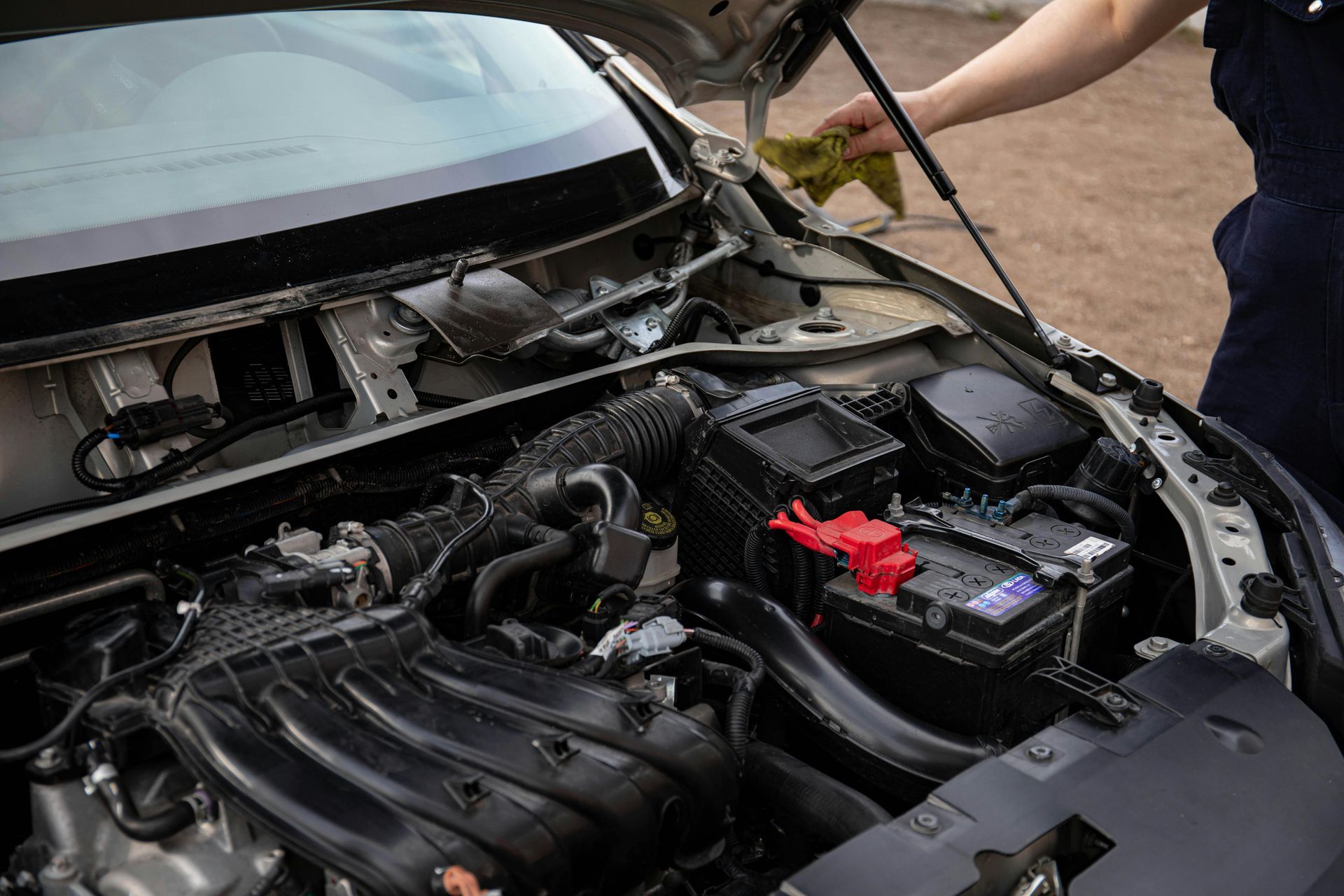 Open car hood with engine bay exposed and a person checking a component near the battery