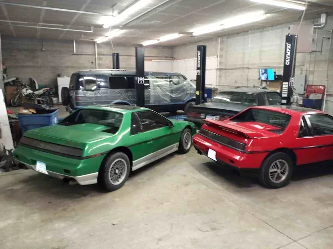 Two sports cars, green and red, parked indoors in a garage with several vehicles in the background