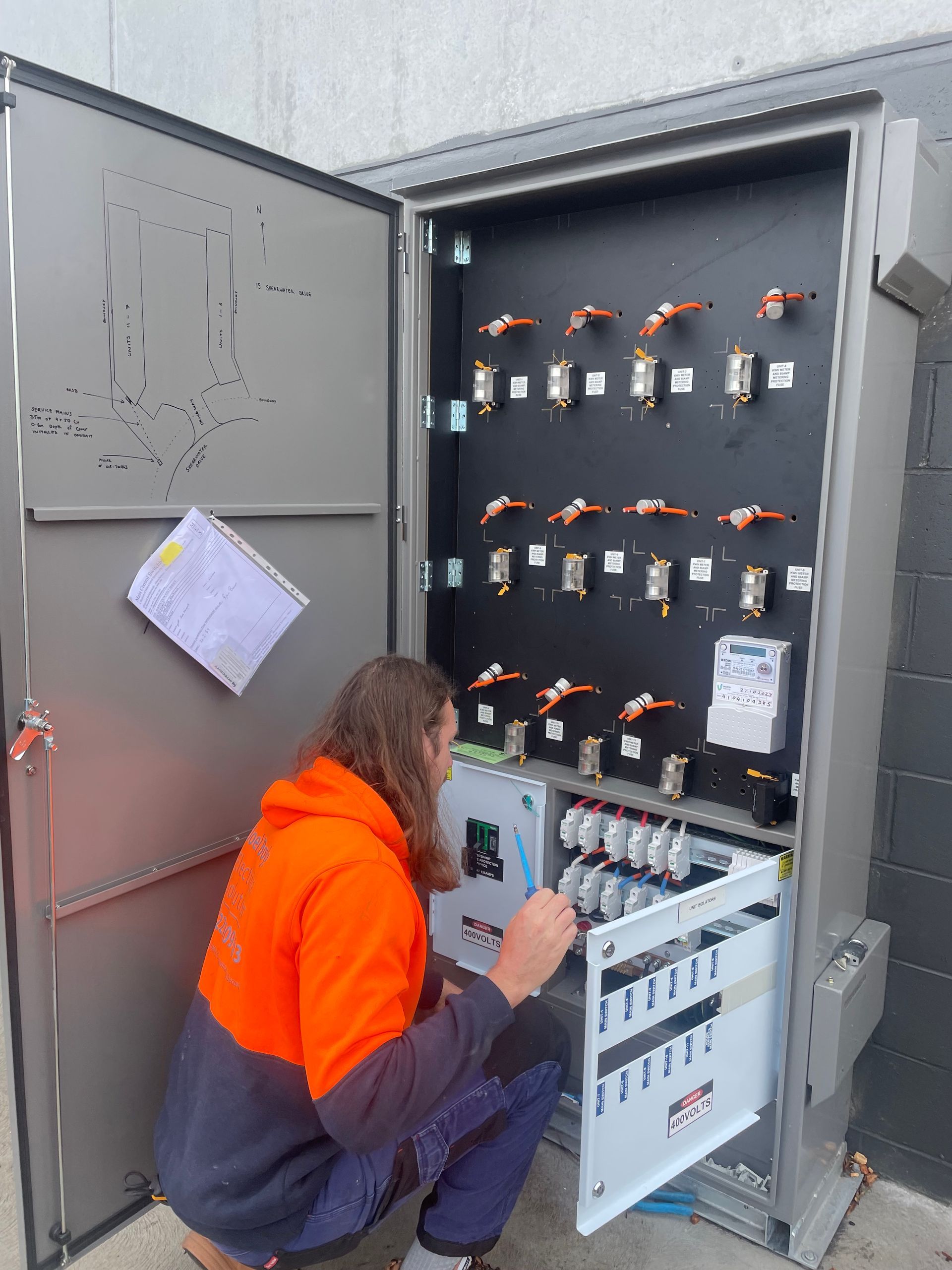 Electrician working inside an open electrical cabinet with switches and meters. Orange and blue work clothes. Outdoors.Powered By JBES In Sandgate, NSW