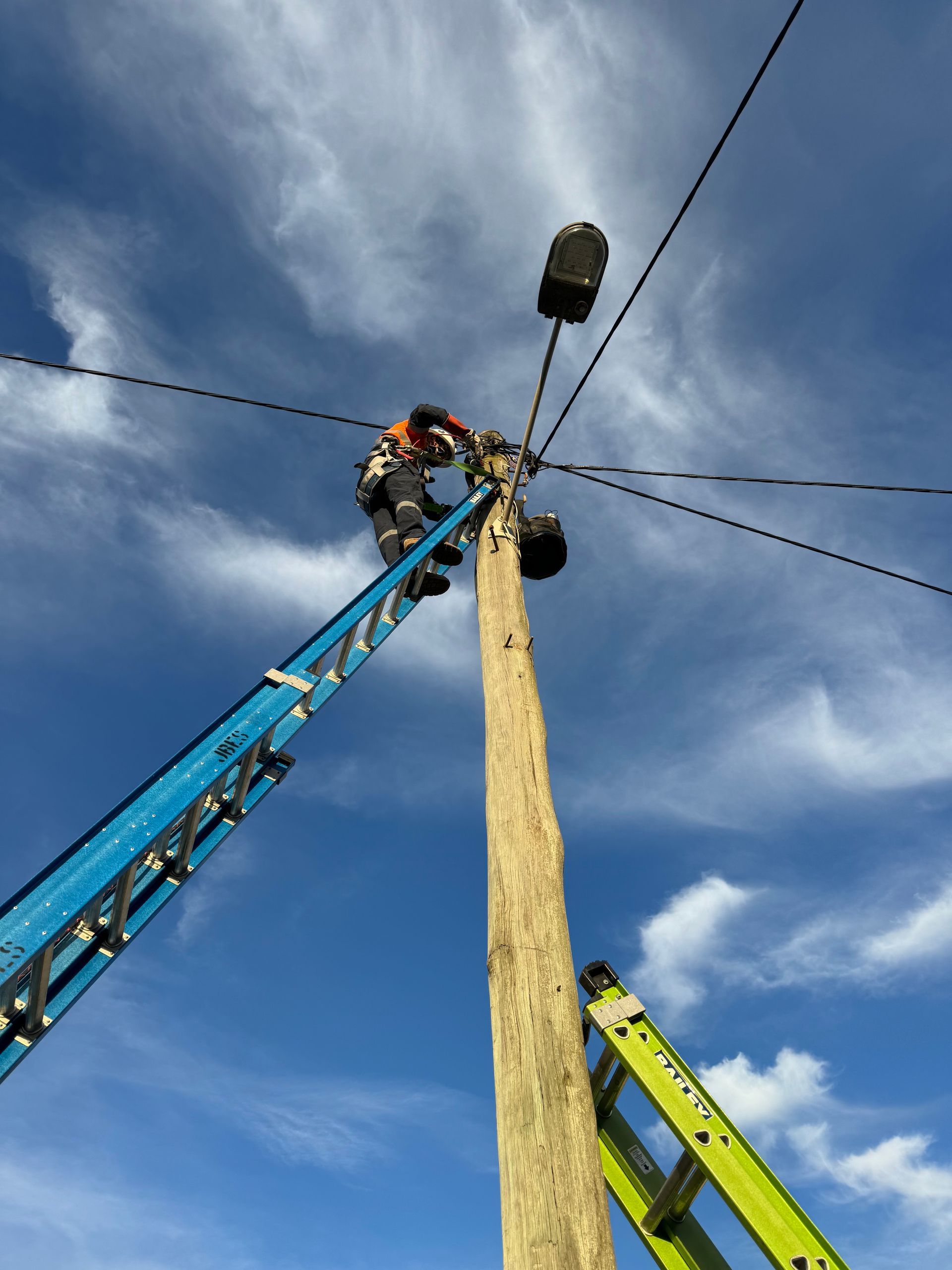Lineman on ladder working on wires atop a utility pole under a blue sky - Powered By JBES In Sandgate, NSW