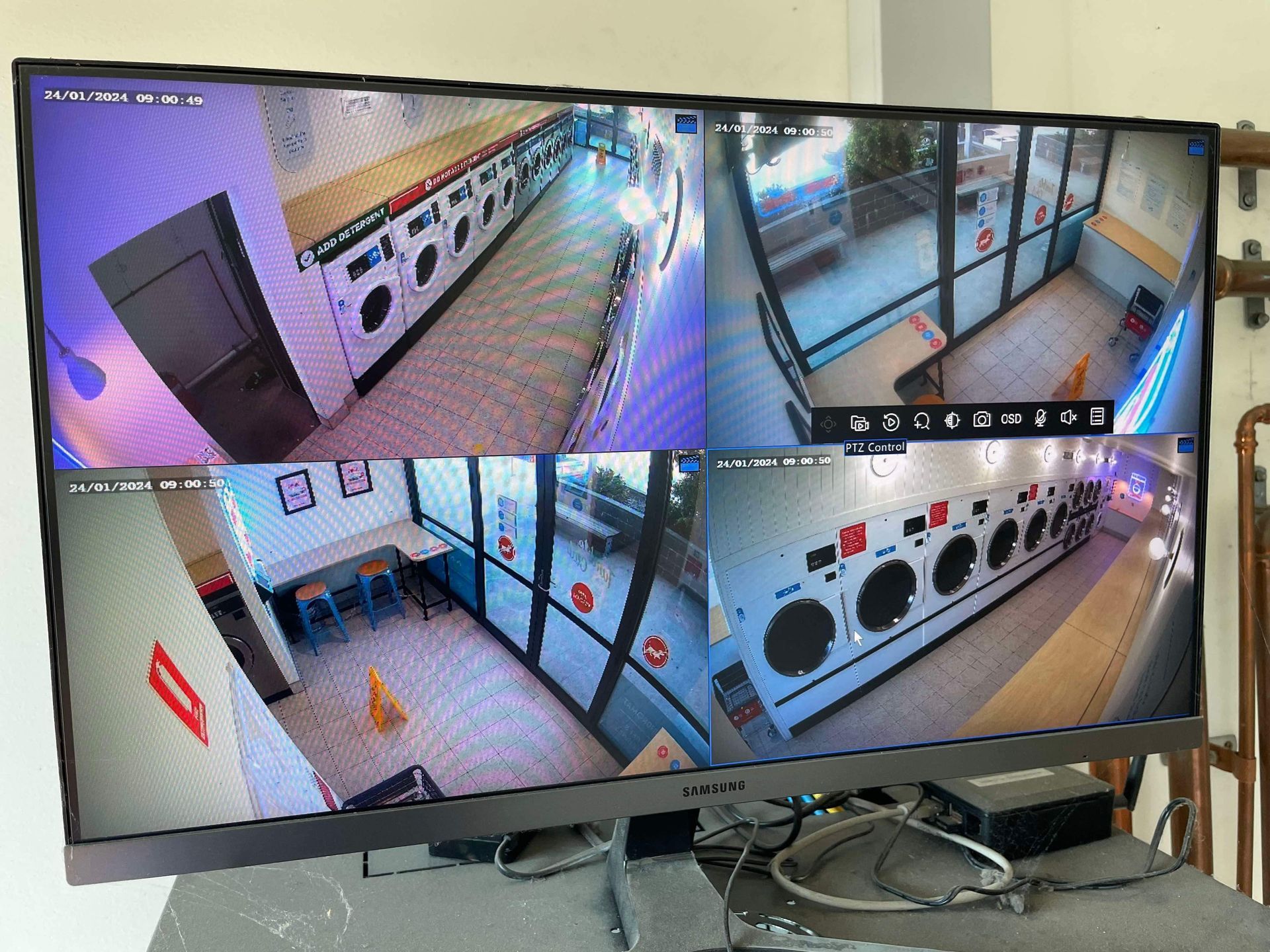 A monitor displaying four camera views of a laundromat: washing machines, tables, and a waiting area.Powered By JBES In Sandgate, NSW