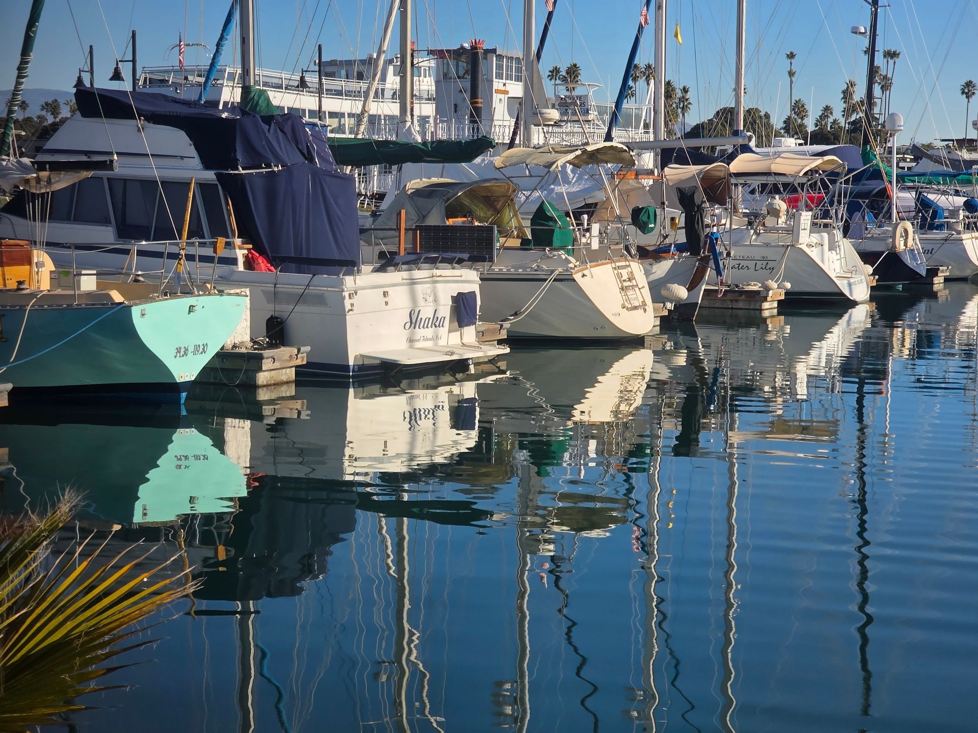 Sailboats docked in a harbor, reflecting in the water. Blue sky and sunny day.
