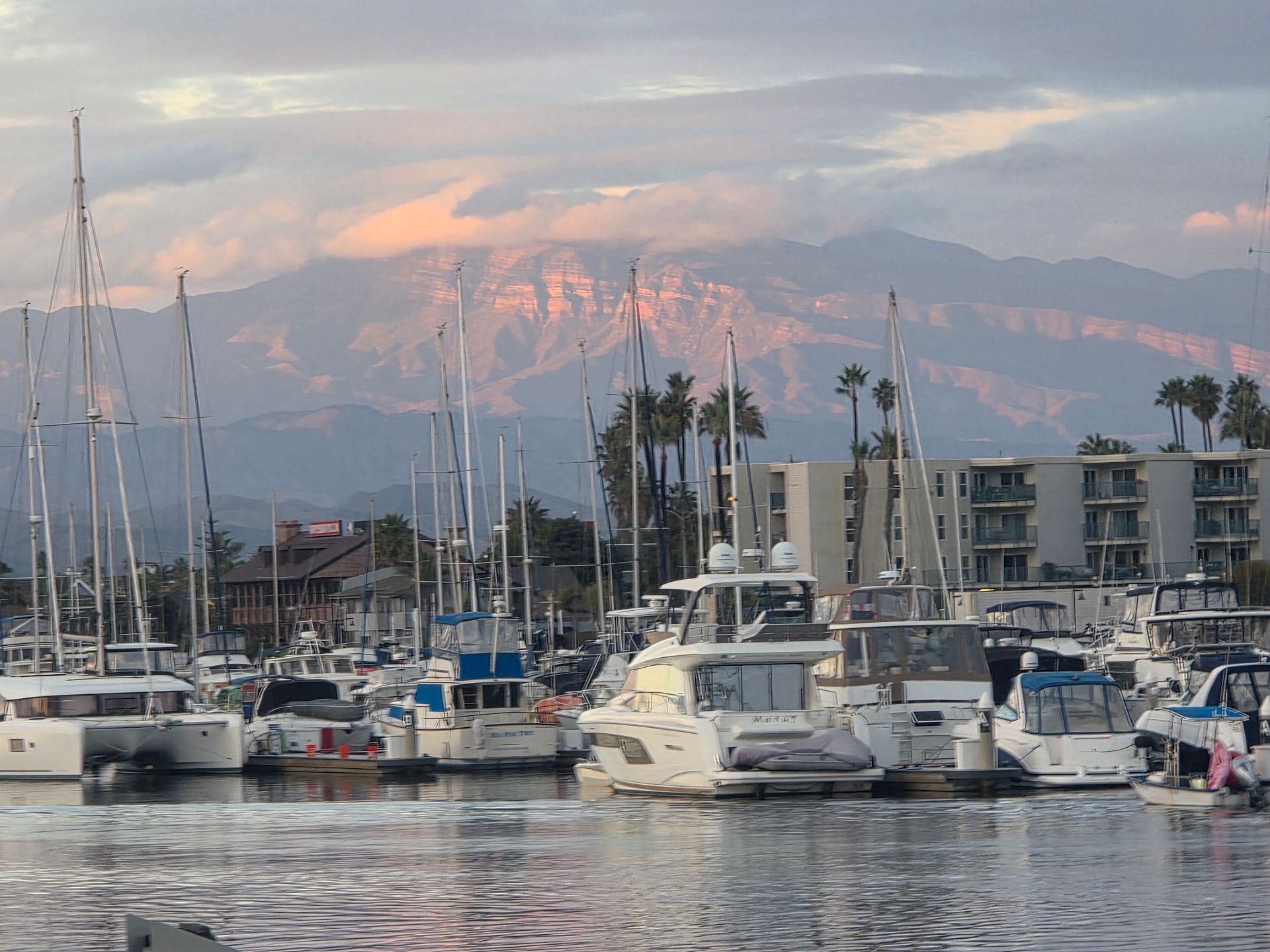 Boats docked in a harbor with a mountain range in the background at sunset, under a cloudy sky.