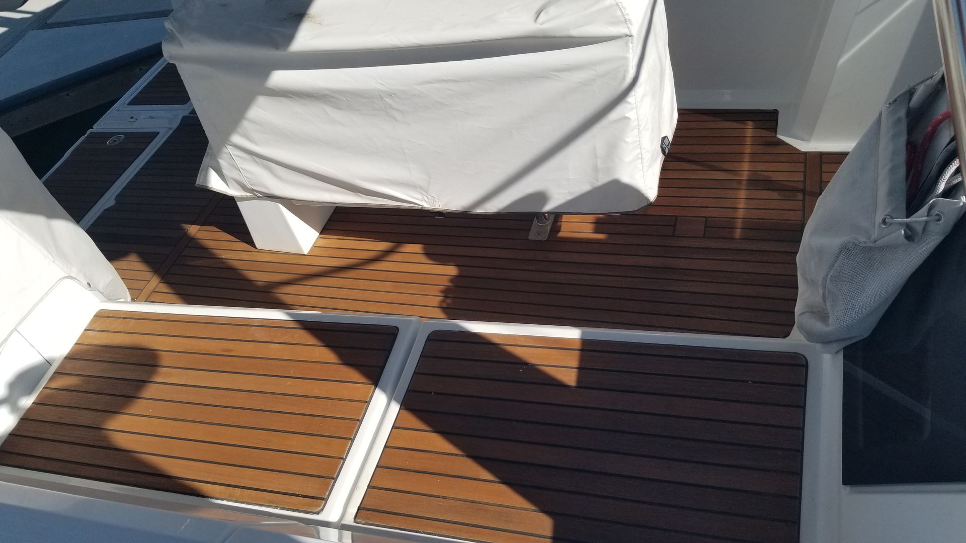 Boat deck with teak-style flooring and white accents. A covered table is centered. Sunlight creates shadows.