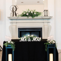 Wedding fireplace mantel with white floral arrangement, black drape, and “Mr & Mrs” sign