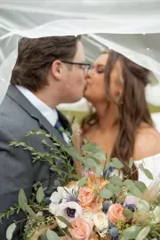 Couple kissing under a veil, holding a bouquet of pastel flowers and greenery