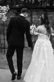 Newlyweds in formalwear walking hand in hand along a garden path.