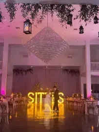 Pink-lit wedding reception with illuminated “SIT” letters, chandeliers, and a couple dancing on the floor