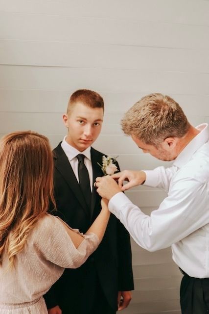 Two people pin a boutonniere on a young man in a black suit, likely before a formal event.