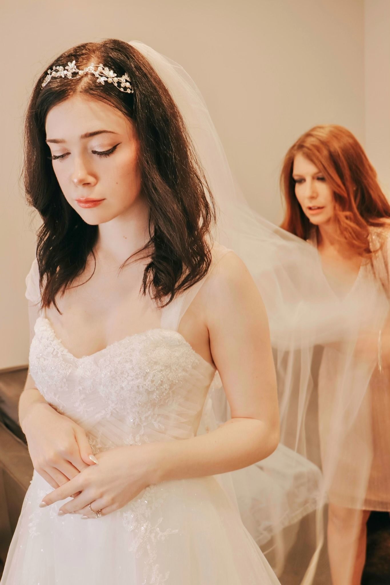 Two women in white dresses and veils stand in a soft-lit bridal scene.