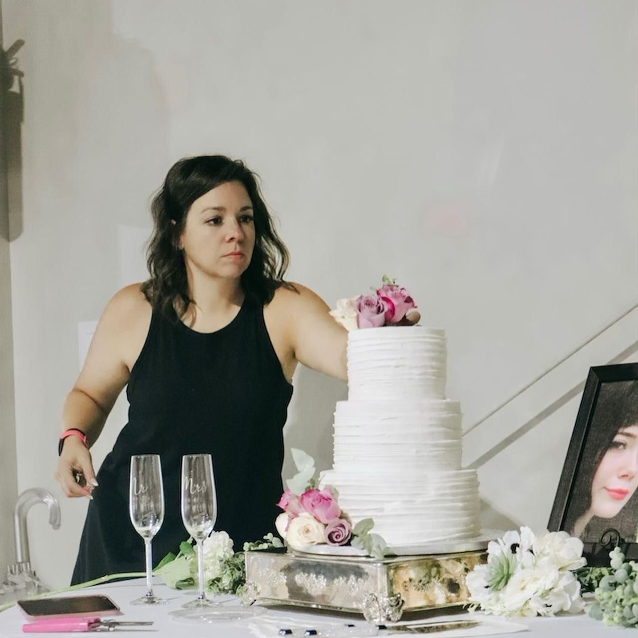 Woman beside a white tiered cake decorated with pink flowers at a party table
