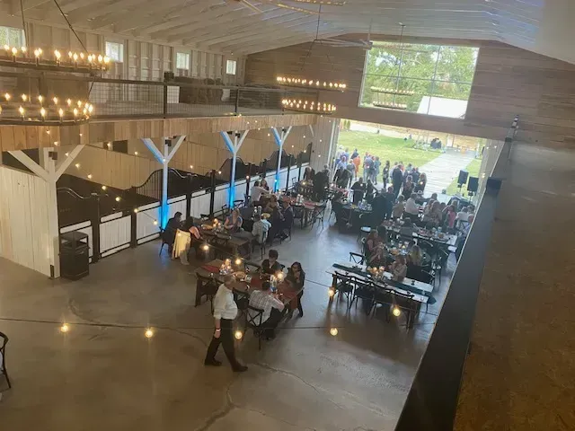 Large rustic barn venue with long tables, seated guests, and warm string lights during a gathering