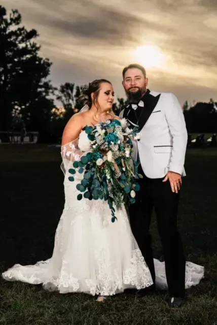 Wedding couple posing outdoors at sunset, bride in white gown holding blue bouquet, groom in white jacket.