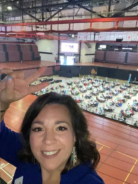 Smiling woman waving from balcony overlooking a large indoor banquet hall with round tables and guests