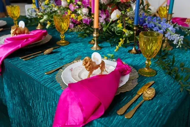 Festive table setting with teal tablecloth, pink napkins, gold cutlery, candles, and floral arrangements