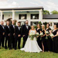 Wedding party posing in formal black attire before a white estate, with bride in a white gown holding a bouquet.