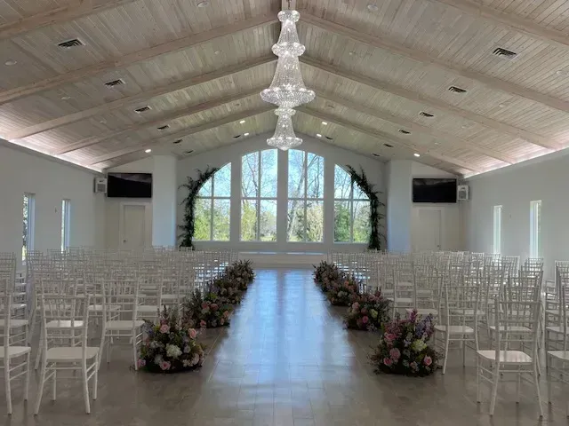 Bright wedding ceremony hall with white chairs, floral aisle arrangements, and large arched windows