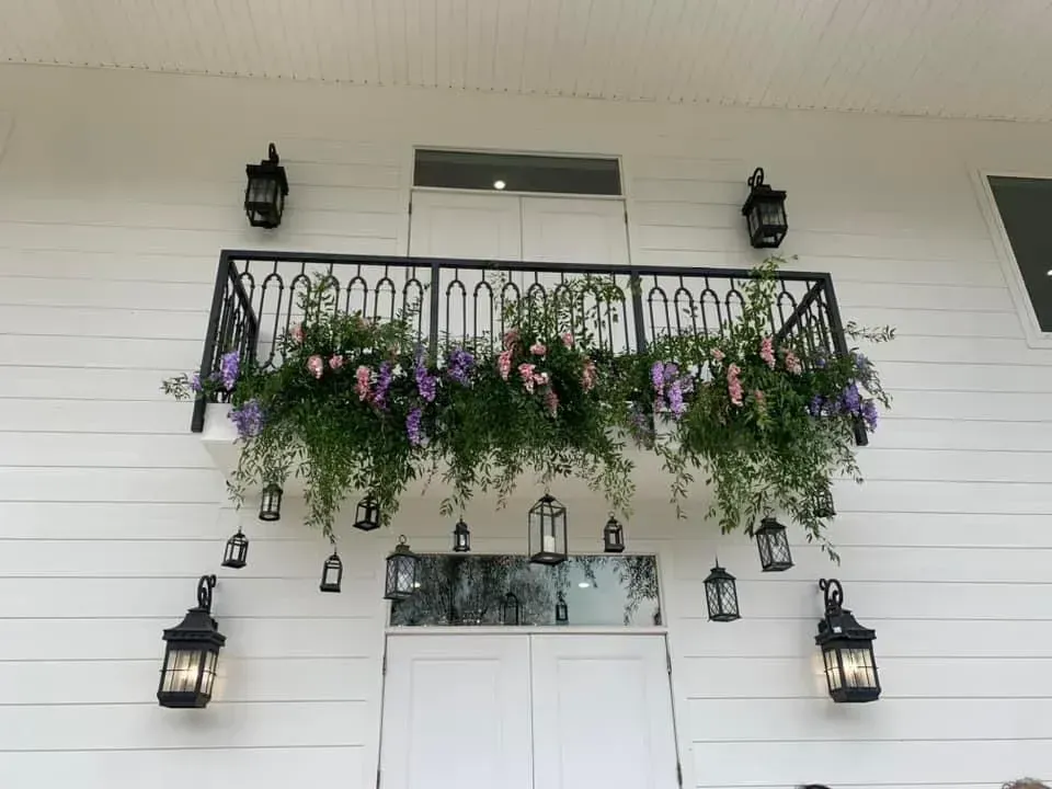 White house facade with black balcony railing and hanging purple-green flower baskets over the front door