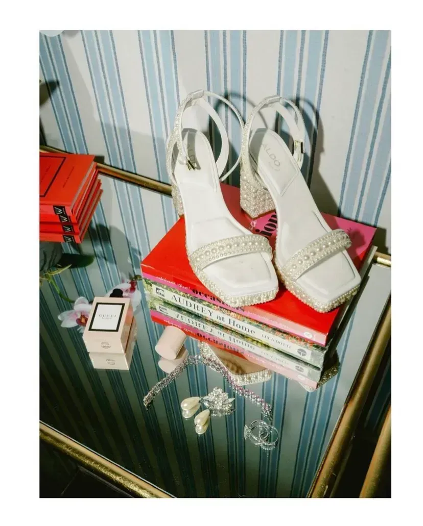 White heeled sandals on stacked books atop a mirrored table beside a red box.