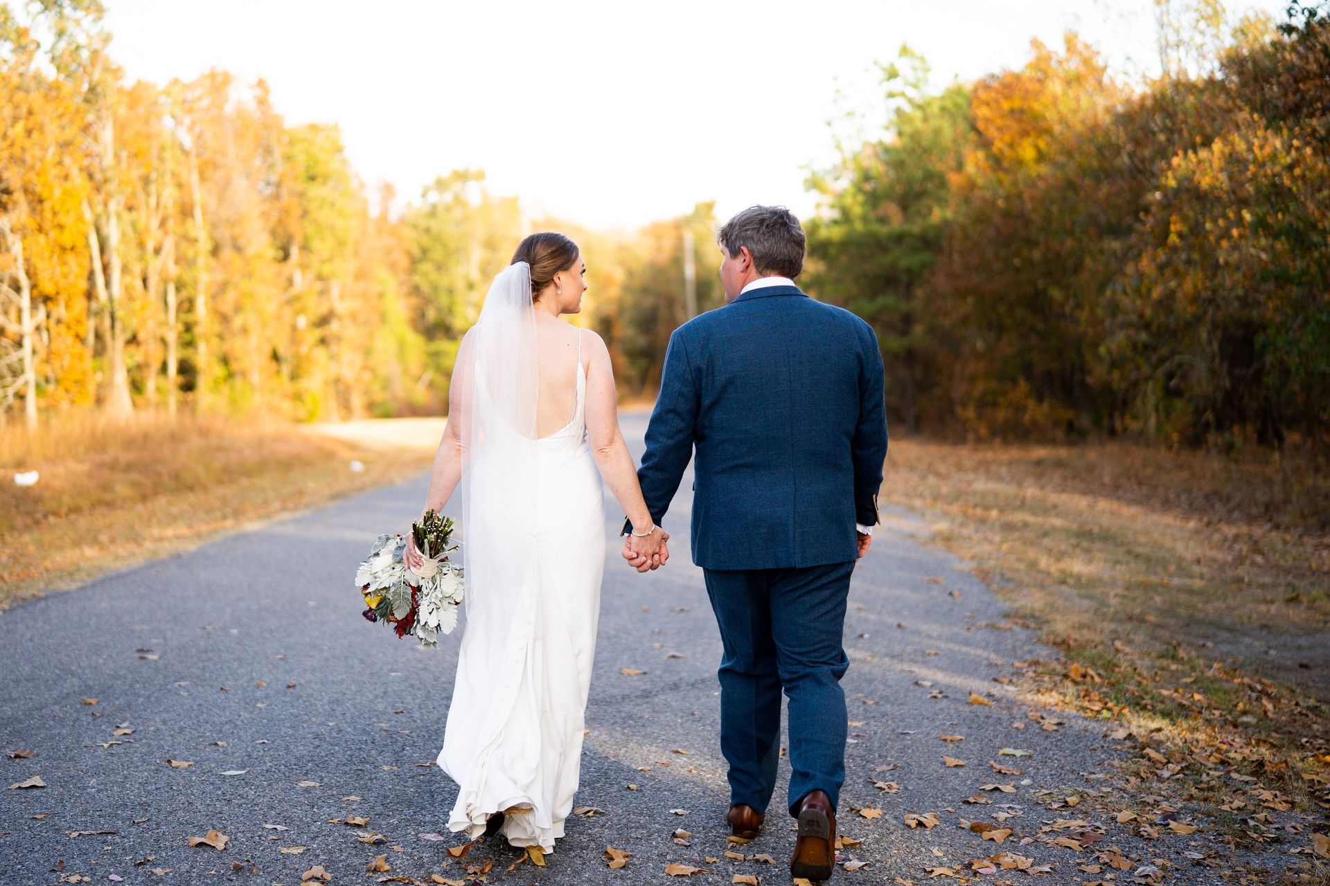 Newlyweds walking hand in hand down a leaf-strewn road, bride in white dress and groom in navy suit
