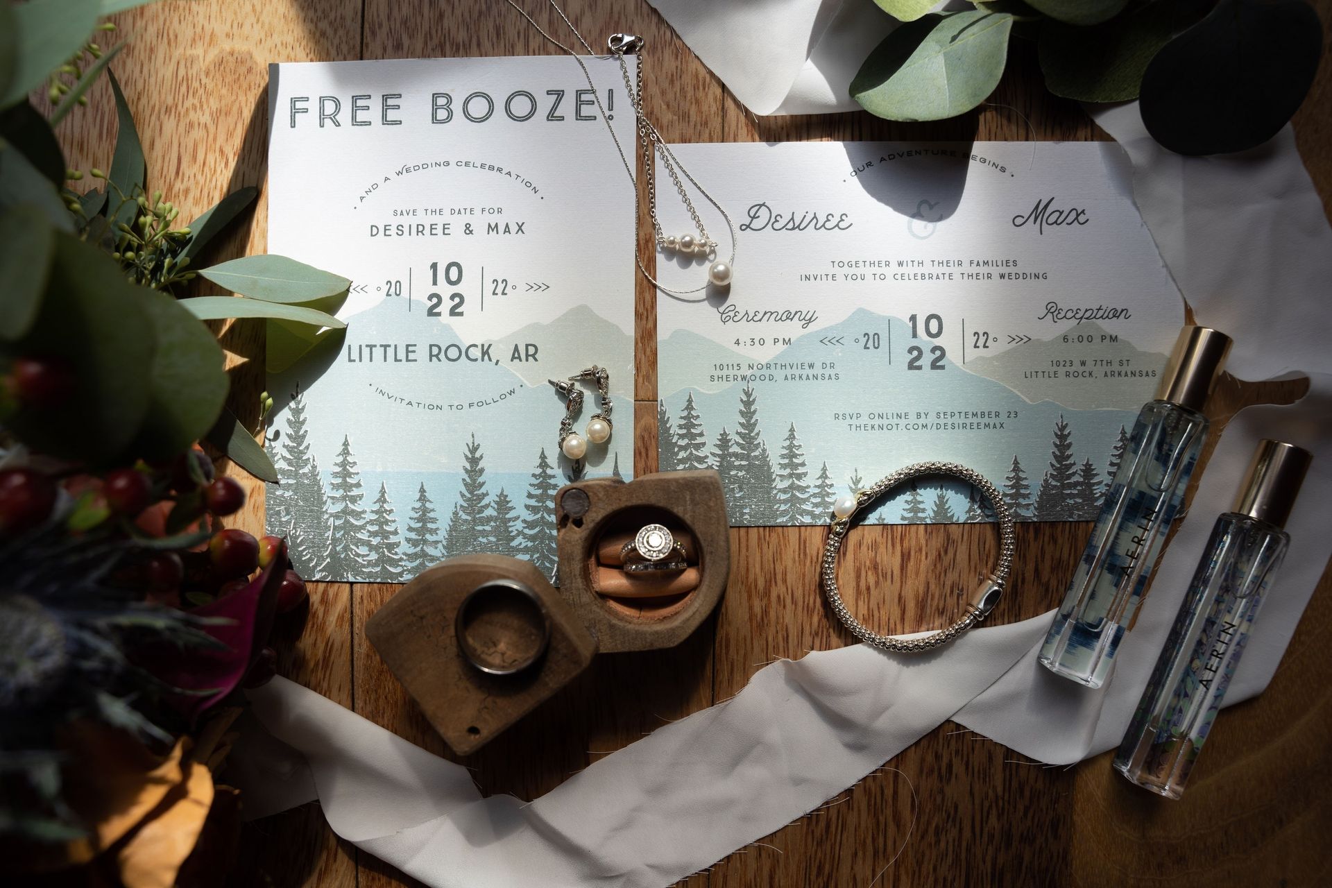 Rustic wedding stationery with “Free Booze!” sign, invitations, rings, and pen on a wooden table