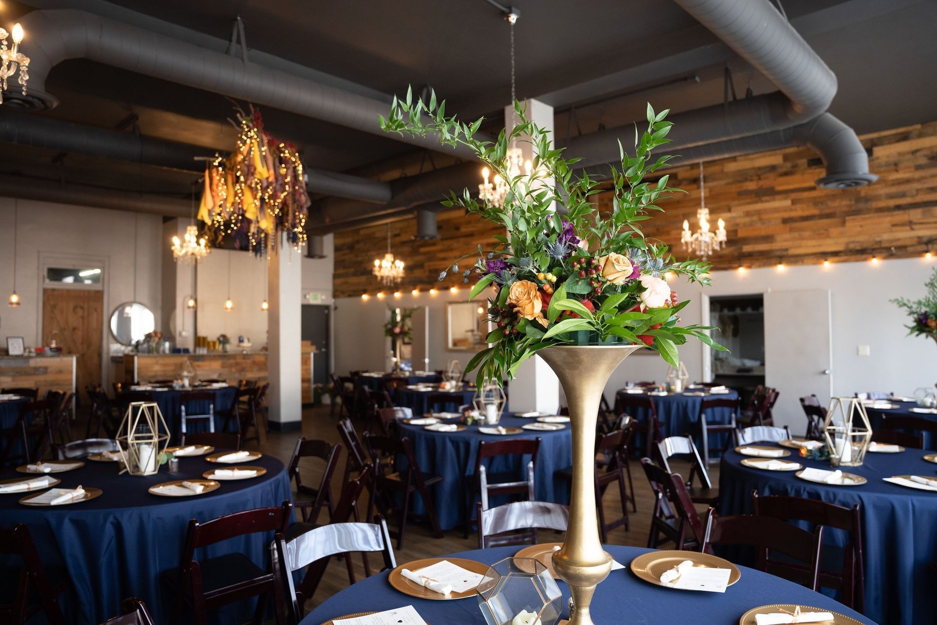 Elegant banquet hall with round navy tables, white chairs, and large floral centerpieces under exposed ceilings