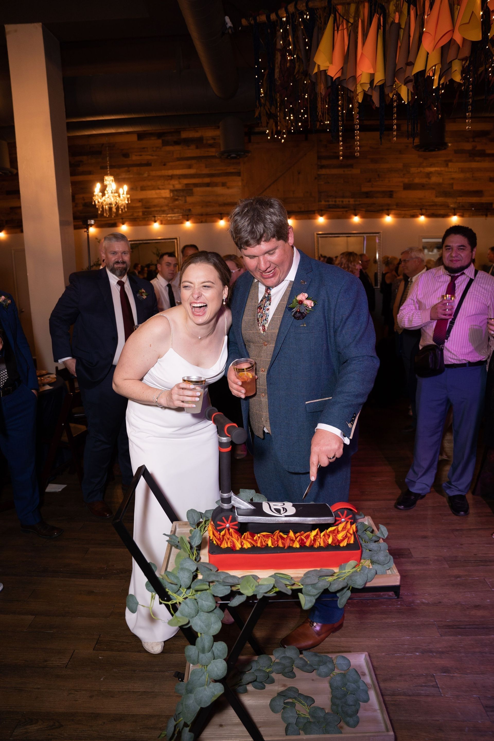 Couple cutting a decorated cake at a wedding reception, smiling and holding a drink
