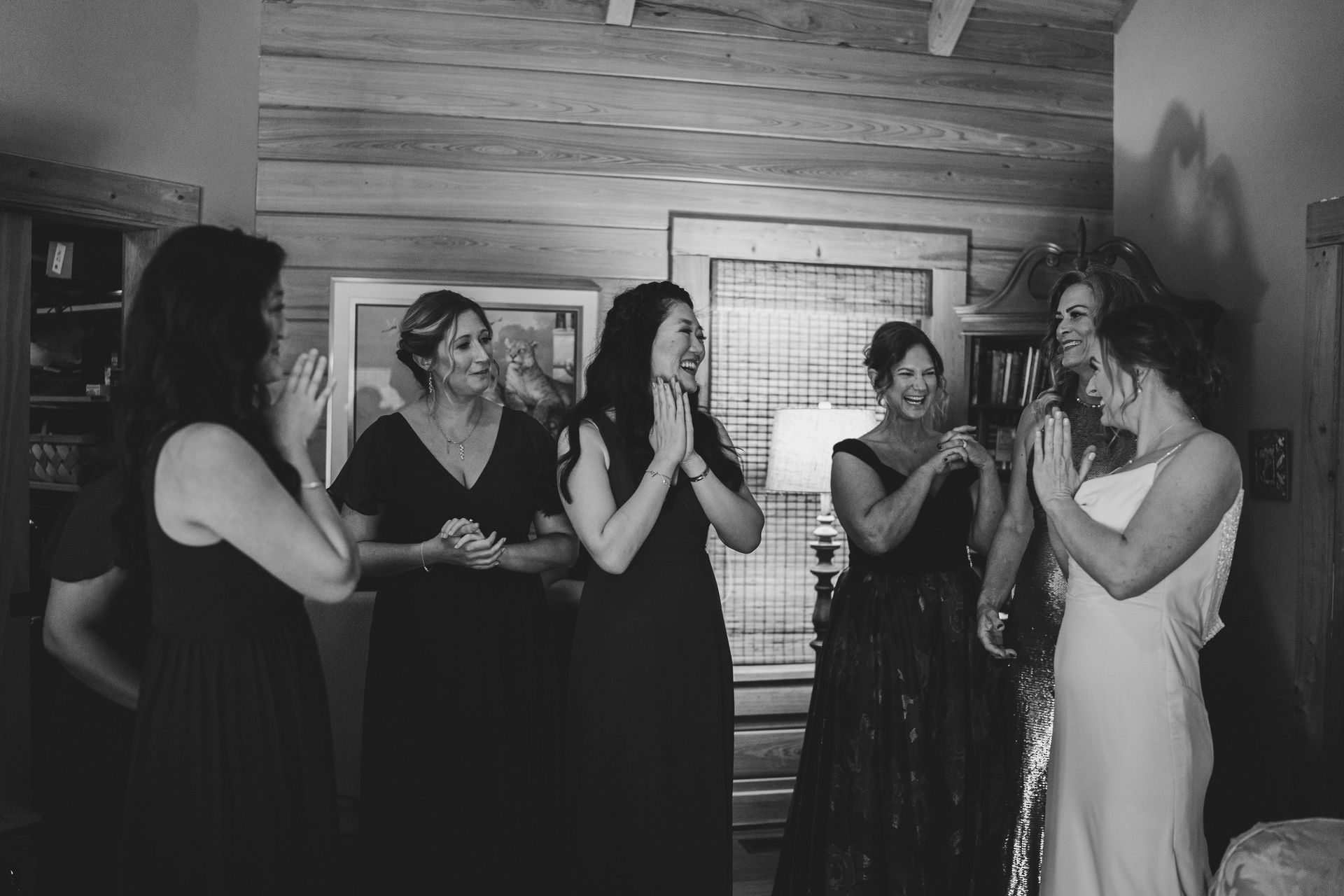Five women in dresses applauding in a rustic indoor setting, one at center with hands clasped.