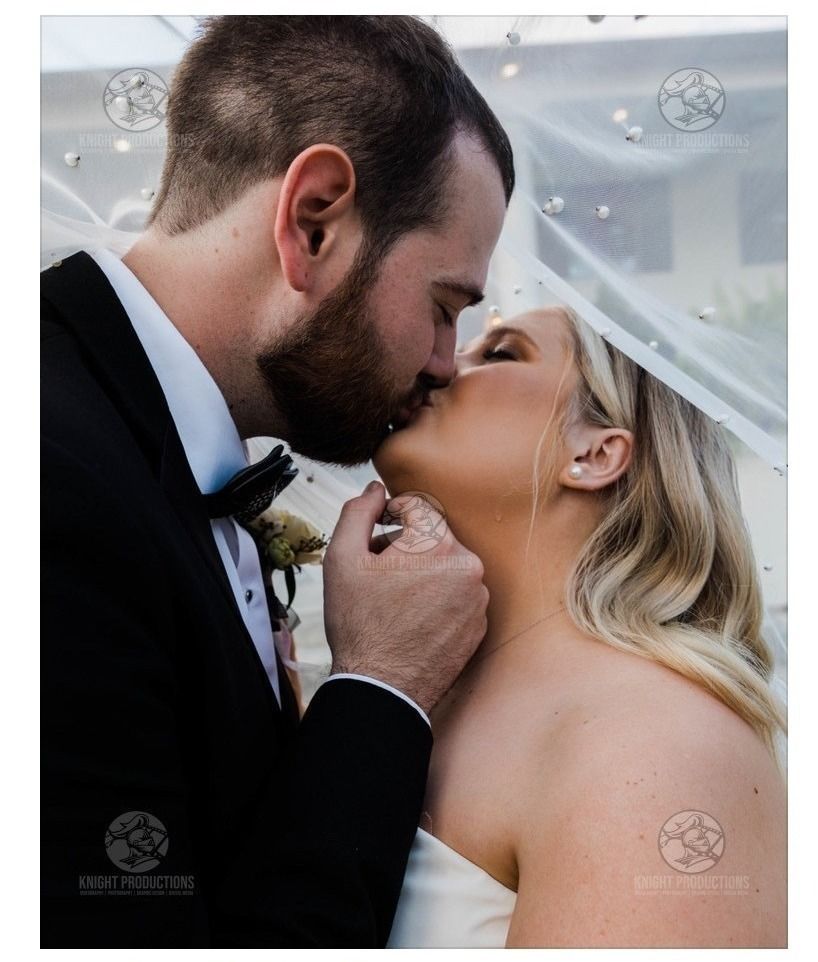 Bride and groom sharing a kiss under a veil, with the groom gently holding her chin.