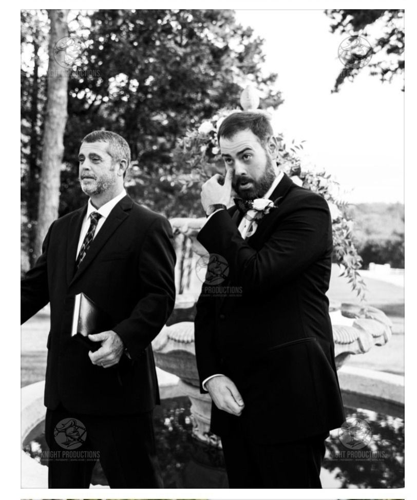 Two men in dark suits standing outdoors at a wedding, one holding a book, with trees and cars behind them.
