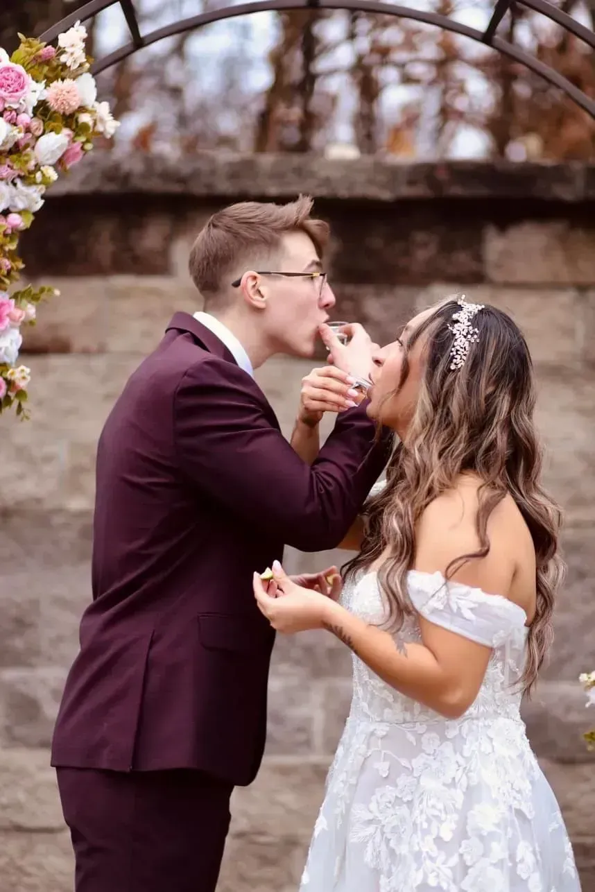 Couple in wedding attire sharing a kiss outdoors beside an arched stone wall and blossoms