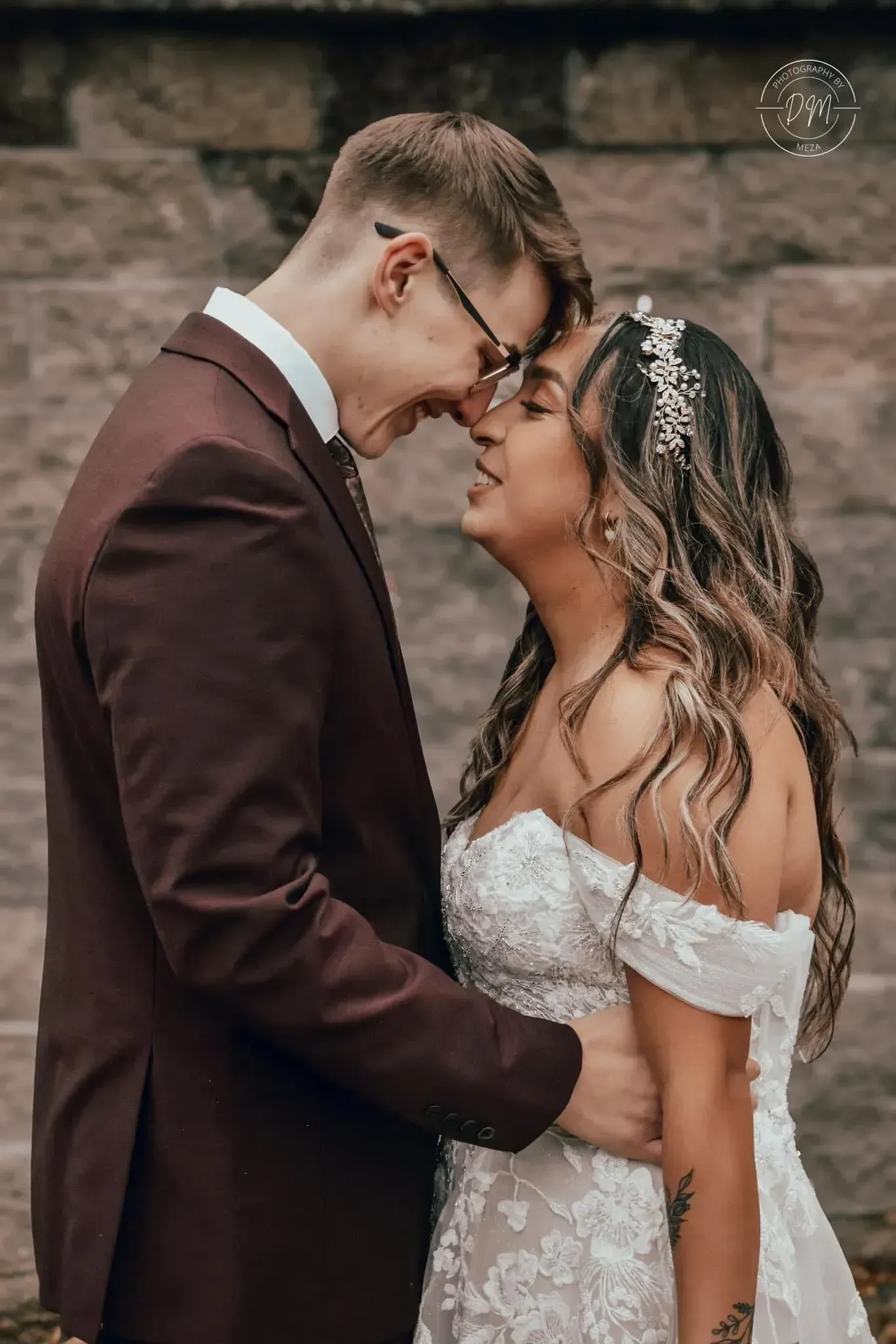 Couple in wedding attire touching foreheads outdoors, bride in white lace dress and groom in maroon suit.