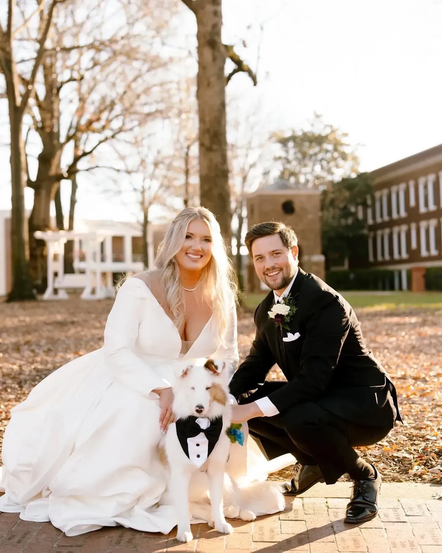 Bride and groom crouch beside a dog outdoors in front of a building.