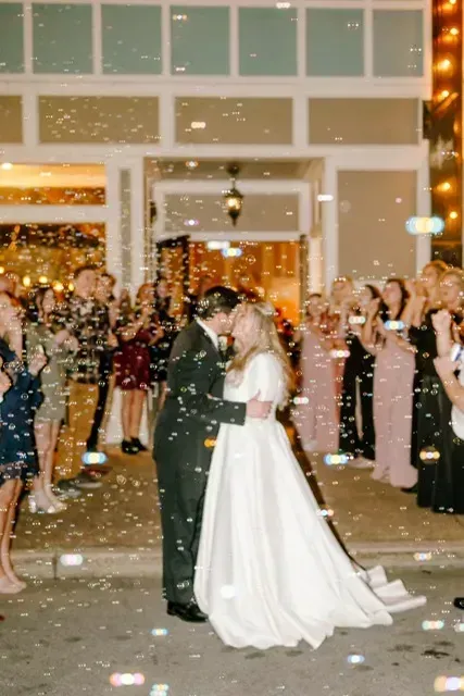 Newlyweds kiss at the end of an outdoor wedding aisle as guests celebrate with confetti