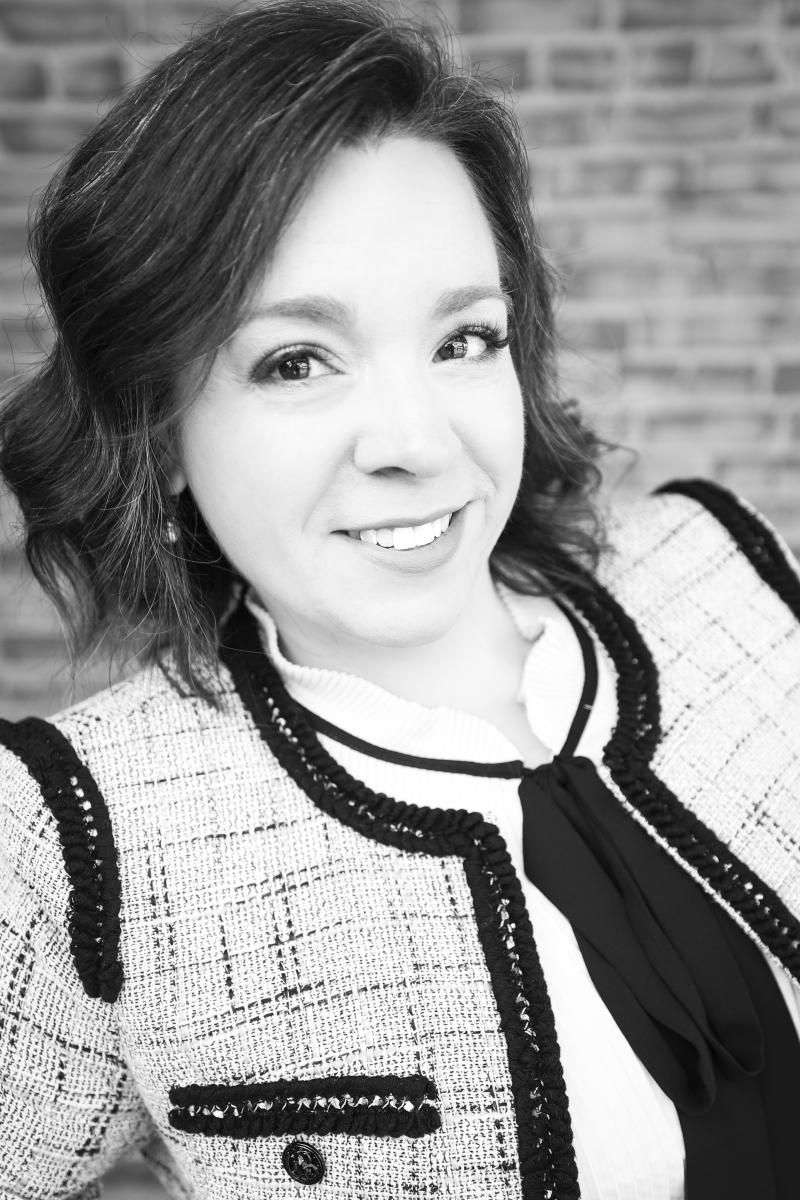 Smiling person in a light tweed jacket with black trim against a brick wall background