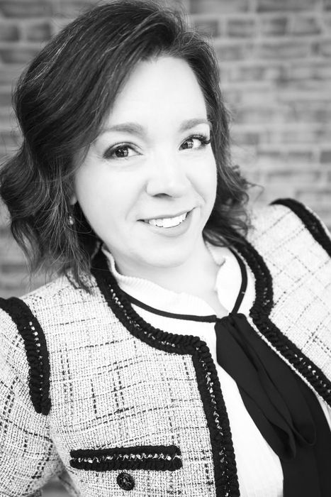 Smiling person in a light tweed jacket with black trim against a brick wall background