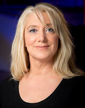 Blonde vrouw met een lichte huid glimlacht, gekleed in een zwart topje tegen een wazig blauwe achtergrond.